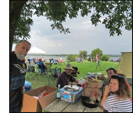 Picture shows a picnic table under a large tree, filled with boxes and bags, jugs of water, etc.  Stephanie and Erick are on the right side and Dick is on the left side, Fred is standing near the tree, all are smiling at us.  Behind them we see lots of lawnchairs and another picnic table, a canopy, a large telescope on a tripod, and people walking around.